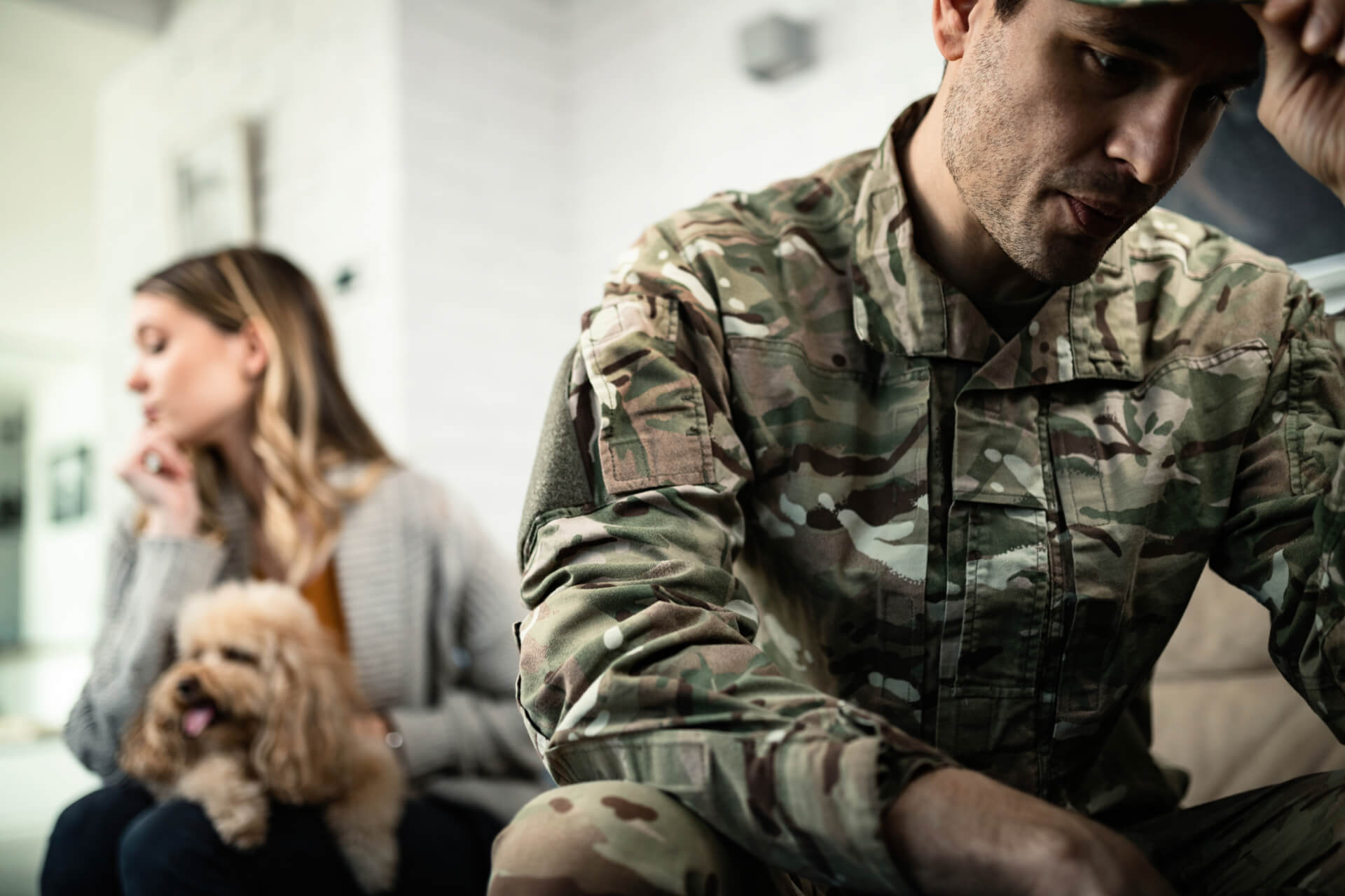 A distressed military servicemember sits in the foreground while his spouse, holding a dog, looks away in the background, representing the emotional challenges of separation and the need for a divorce attorney in Irvine.