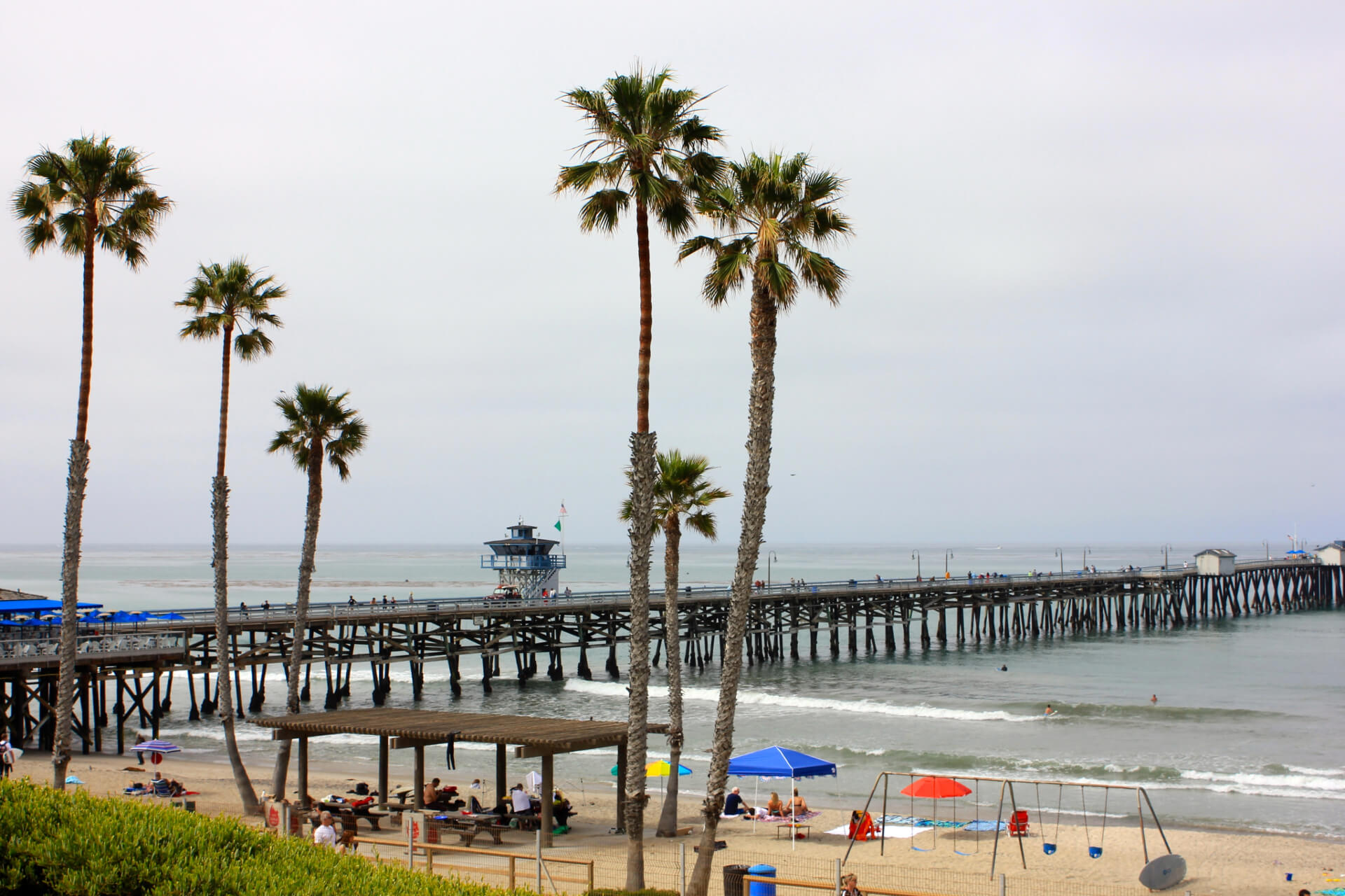 A scenic view of the San Clemente Pier with palm trees, a sandy beach, and people enjoying the ocean, representing the area served by a San Clemente divorce lawyer.